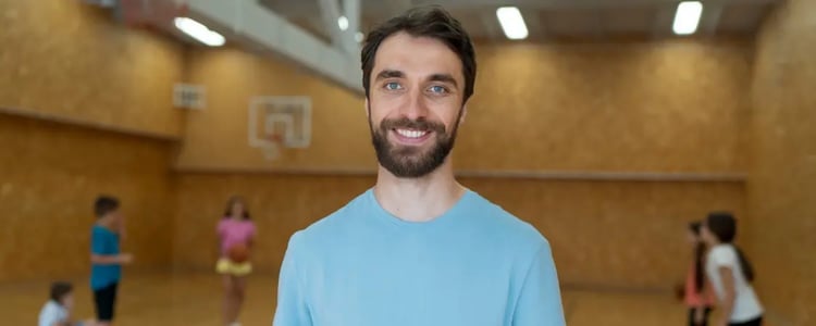 Profesor de educación física sostiene una tabla con clip y hojas mientras supervisa a un grupo de niños que se pasan un balón de básquetbol